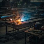 Welding sparks fly as a welder joins metal beams on a workbench; protective helmet and gloves sit nearby.