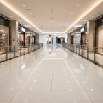 Bright, modern shopping mall corridor with glass storefronts and a glossy tiled floor; empty path lined with railings on both sides at center.”] ,