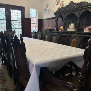 Long, dark carved dining table with matching ornate chairs in a vintage dining room; china and plates on a carved sideboard.